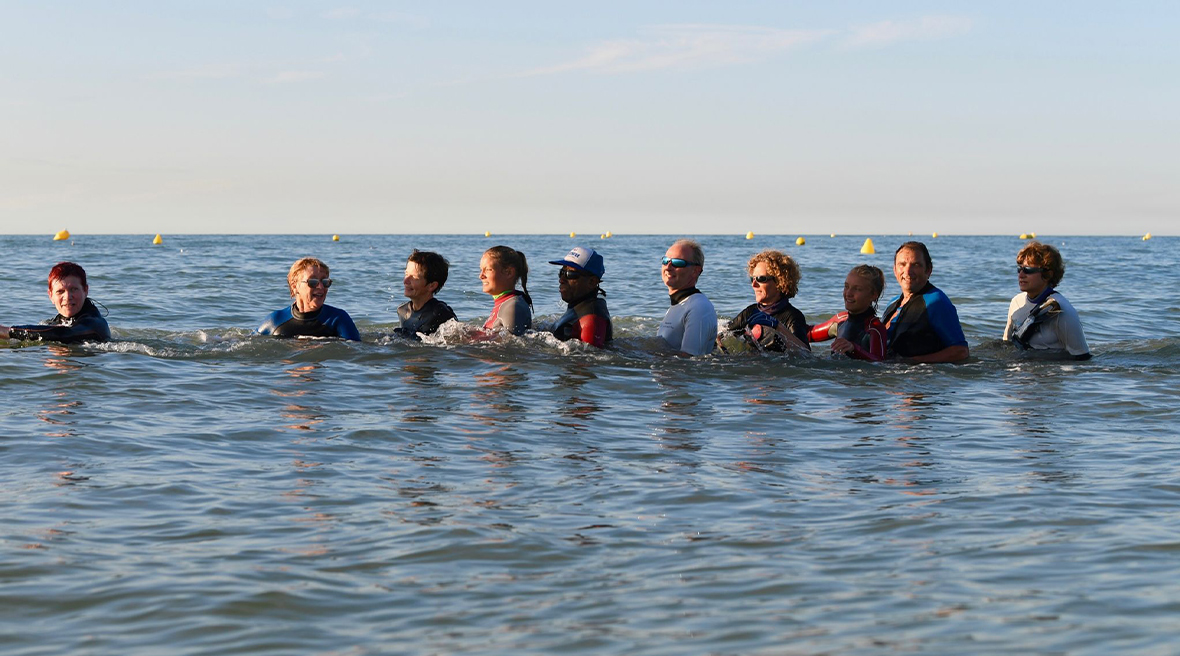 A group of people walk in the sea in a group, wearing wetsuits and wading at waist depth